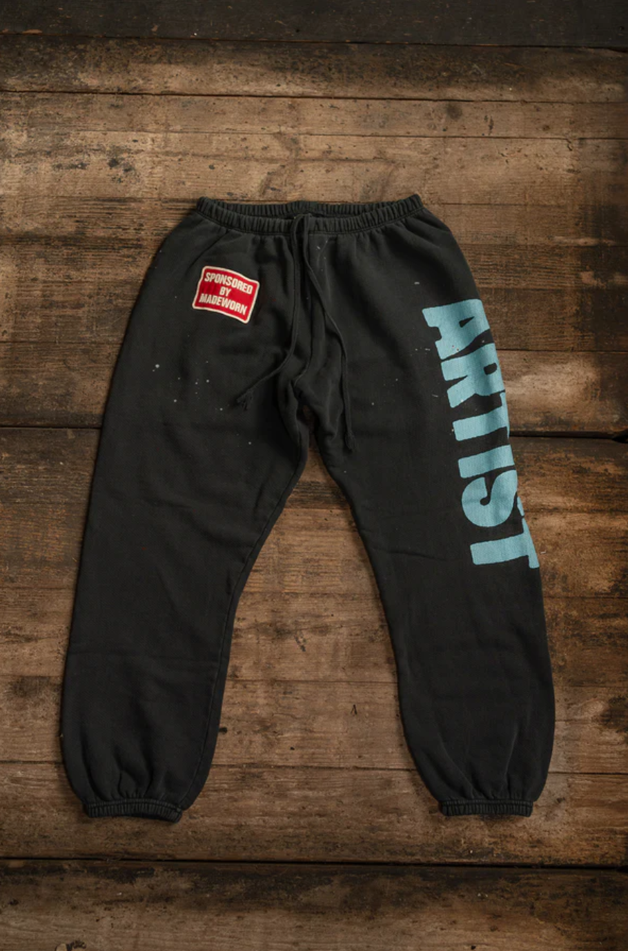 The MadeWorn P.E. Sweatpants by Made Worn are unisex black sweatpants made from heavyweight French terry cotton. They feature "ARTIST" in bold blue letters on the right leg and a small red "SPRAYED GOOD HANDWORK" patch on the left hip. Designed in vintage style, they are crafted in Los Angeles.