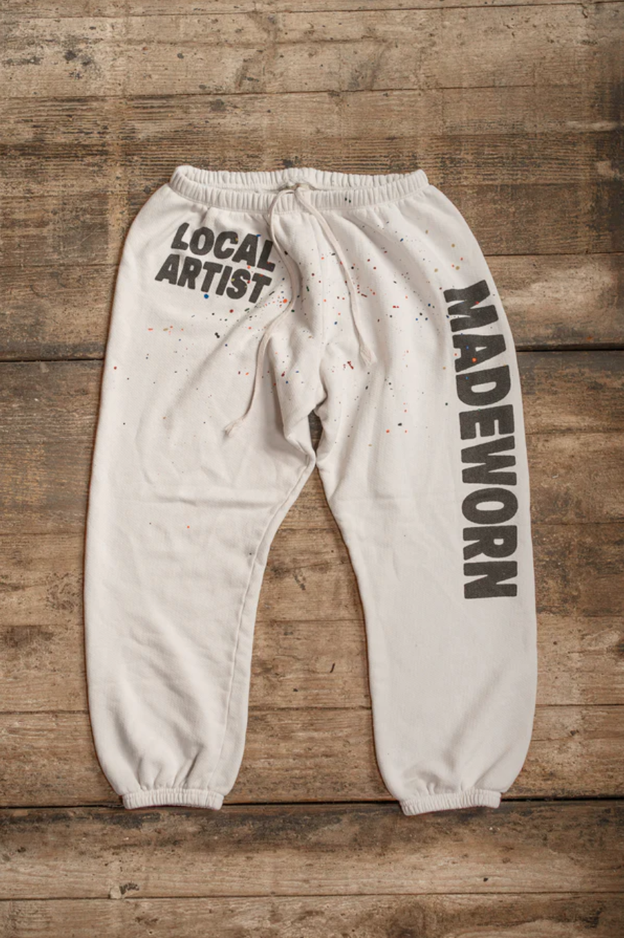 The PE Sweatpant by Made Worn, crafted from soft French Terry Cotton and designed with "LOCAL ARTIST" on the left leg and "MADEWORN" on the right, features small paint splatters for an artistic touch. Displayed on rustic wood, these vintage white sweatpants highlight their Made in USA quality.