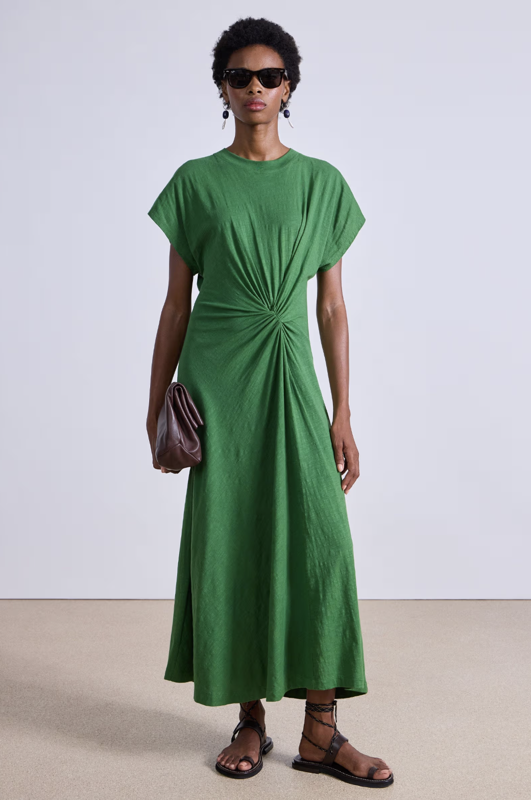 A person wears the Apiece Apart Volta Twist Dress in green organic cotton, featuring short sleeves and a waist twist detail. They accessorize with sunglasses, hoop earrings, and a brown clutch, completing the look with brown strappy sandals.