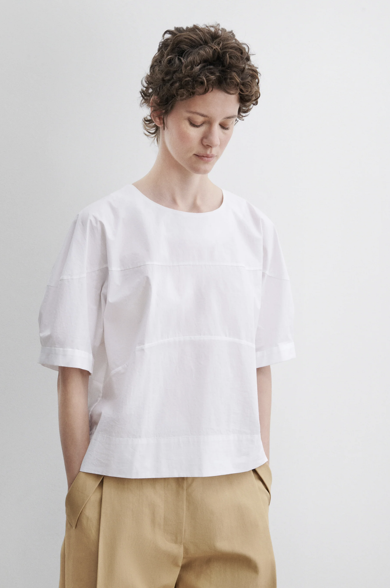 A person with short curly hair wears the Rachel Comey Rugby Top—a loose, boxy short-sleeve piece with a crew neckline—paired with tan pants, standing against a plain light background and looking down calmly.