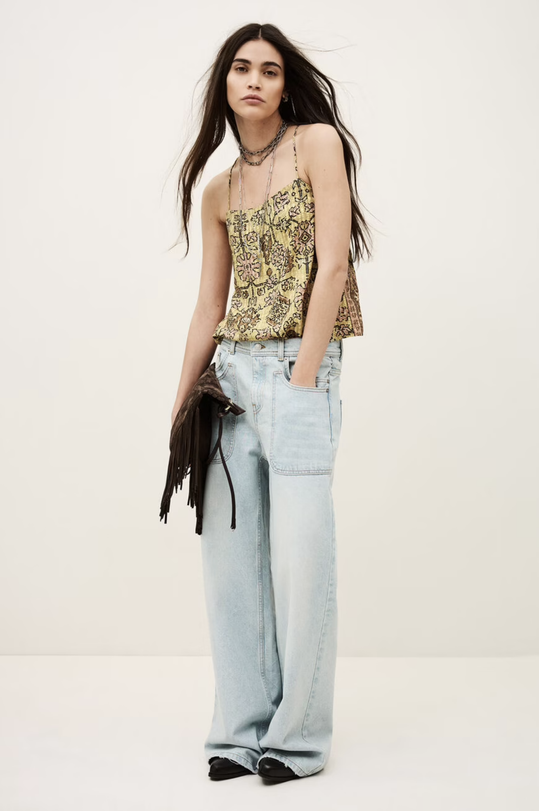 A woman with long dark hair wears the BA&SH Top Pamela, light blue wide-leg jeans, black pointed shoes, layered necklaces, and holds a fringed black bag against a plain light background.