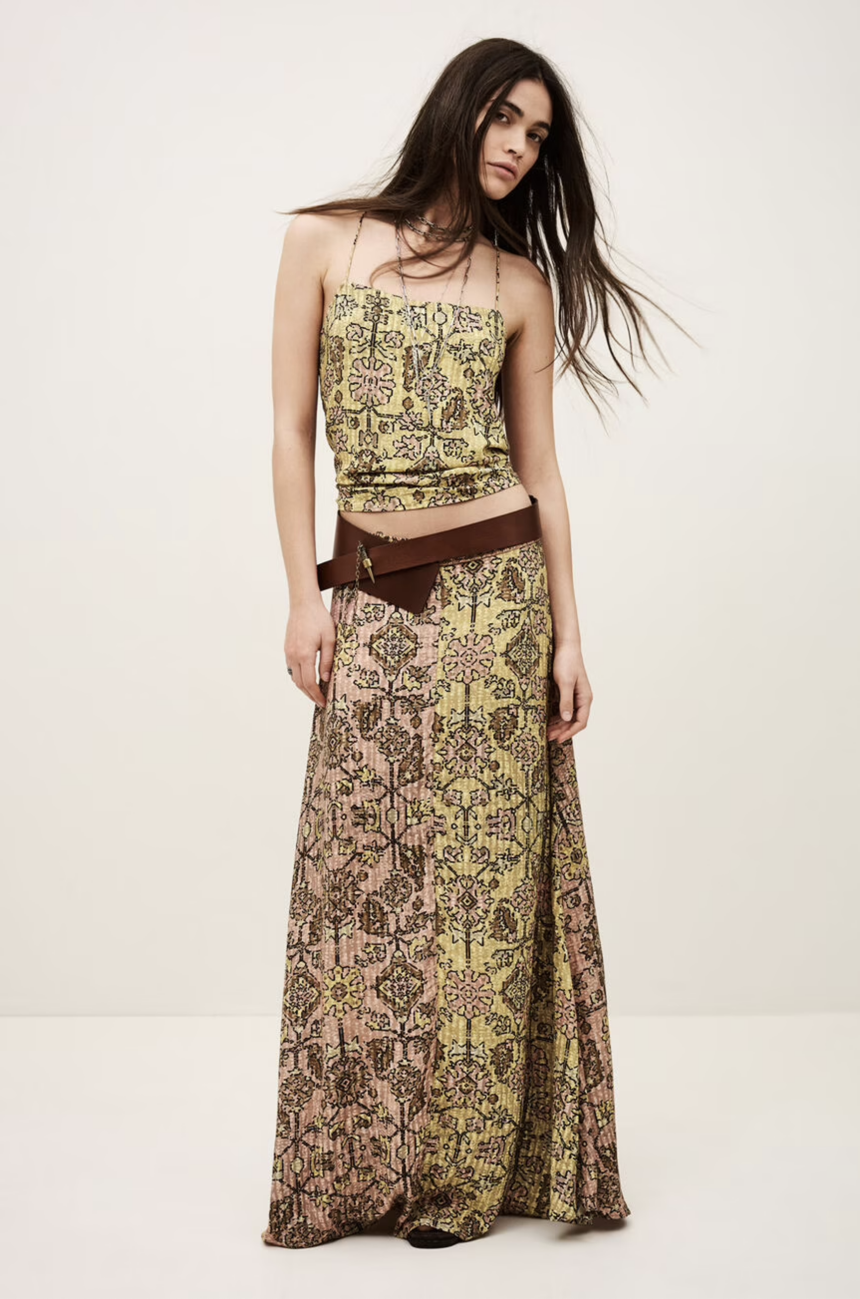 A woman poses against a plain background wearing the BA&SH Skirt Pario—a patterned yellow and brown maxi skirt with a high waist, styled with a wide brown belt. Her long dark hair is worn loose and slightly tousled.