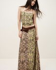 A woman poses against a plain background wearing the BA&SH Skirt Pario—a patterned yellow and brown maxi skirt with a high waist, styled with a wide brown belt. Her long dark hair is worn loose and slightly tousled.