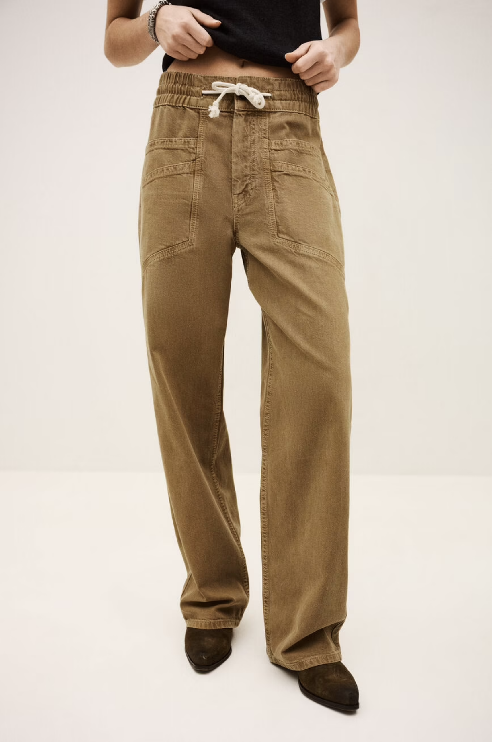 Someone is shown from the shoulders down wearing BA&SH Pant Fima—loose-fitting, light brown cotton linen pants with an elasticated waist and large front pockets—paired with dark pointed shoes, set against a plain background.