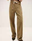 Someone is shown from the shoulders down wearing BA&SH Pant Fima—loose-fitting, light brown cotton linen pants with an elasticated waist and large front pockets—paired with dark pointed shoes, set against a plain background.