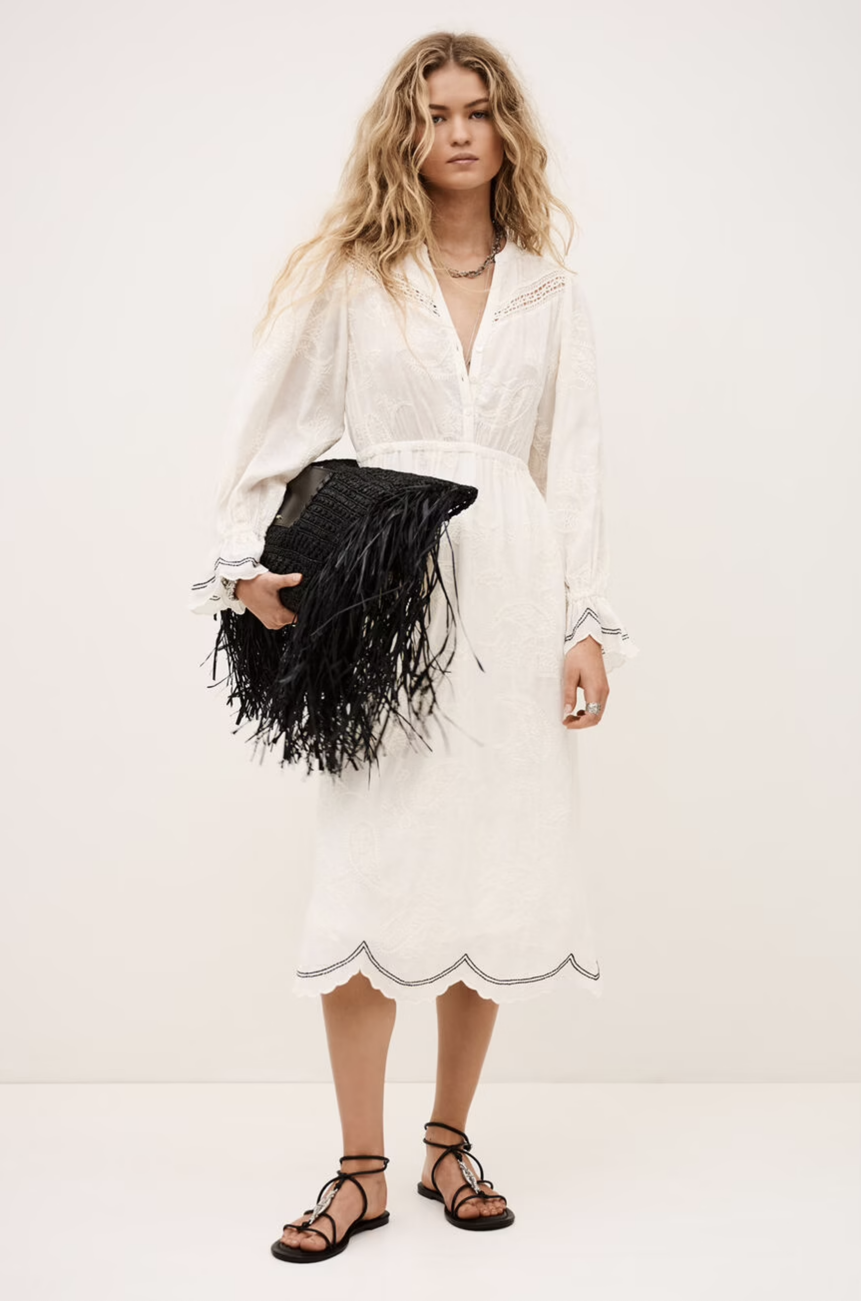 A woman in the BA&SH Dress Prisco, a white long-sleeve lace detail dress, holds a large black fringed clutch. She has wavy blonde hair, wears black strappy sandals, and stands against a plain light background.