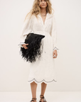 A woman in the BA&SH Dress Prisco, a white long-sleeve lace detail dress, holds a large black fringed clutch. She has wavy blonde hair, wears black strappy sandals, and stands against a plain light background.