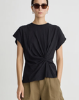 A person wearing sunglasses, silver hoop earrings, the Apiece Apart Volta Twist Top, and light brown shorts stands facing forward against a plain light background.
