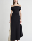 A woman wears the Apiece Apart Marles Off the Shoulder Dress in black cotton jersey with black sandals, a black headband, drop earrings, and holds a cream mesh tote bag, standing against a plain light background.