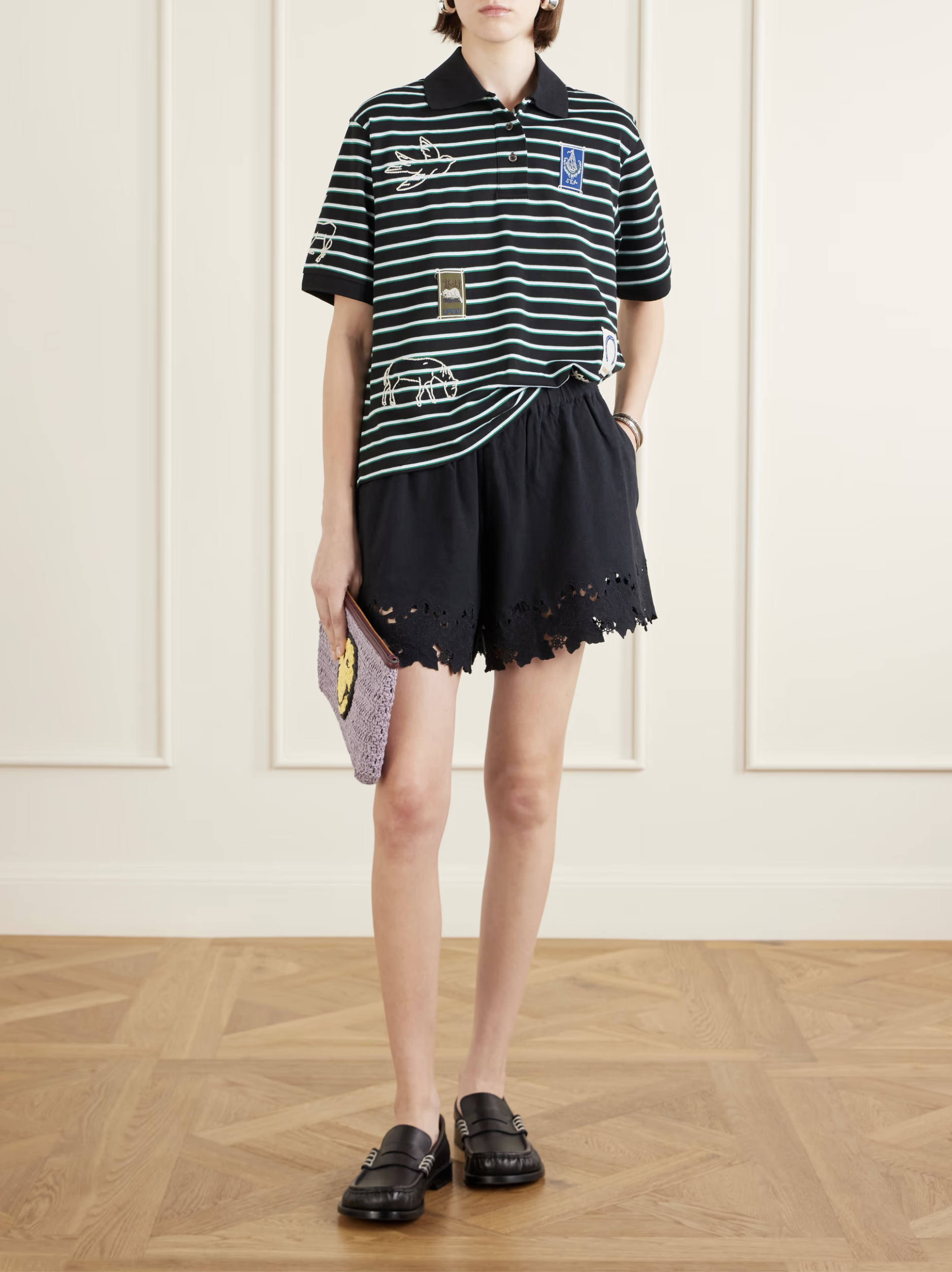 Wearing Sea New York’s Eleannora Shorts, a person indoors pairs them with a green and white striped polo, black loafers, and carries a lavender clutch. The background has white paneled walls and a wooden floor.