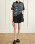 Wearing Sea New York’s Eleannora Shorts, a person indoors pairs them with a green and white striped polo, black loafers, and carries a lavender clutch. The background has white paneled walls and a wooden floor.