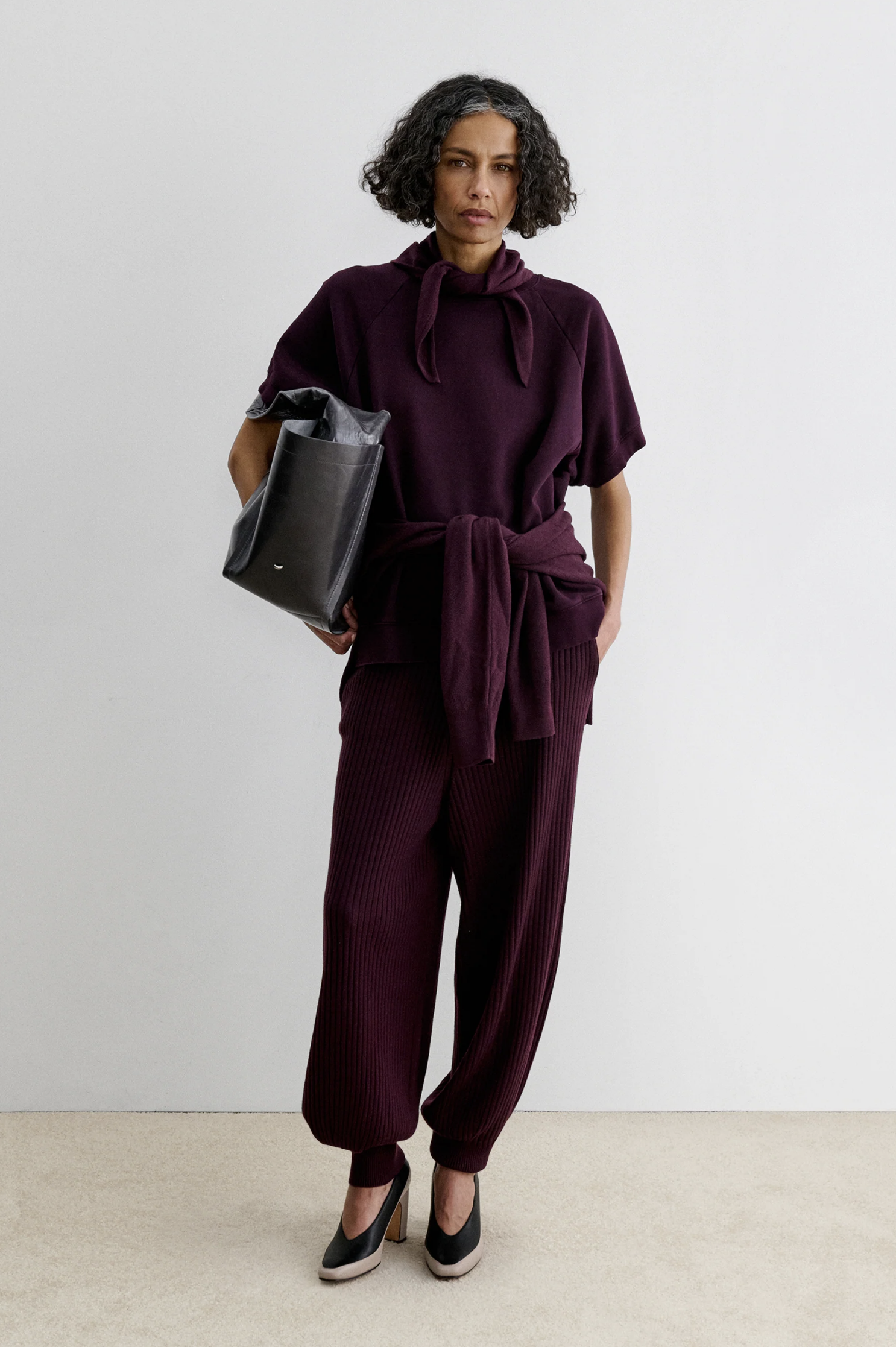 Rachel Comey Nim Pant in burgundy, high-waisted and relaxed fit, styled with a black bag against a white wall.