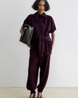 Rachel Comey Nim Pant in burgundy, high-waisted and relaxed fit, styled with a black bag against a white wall.