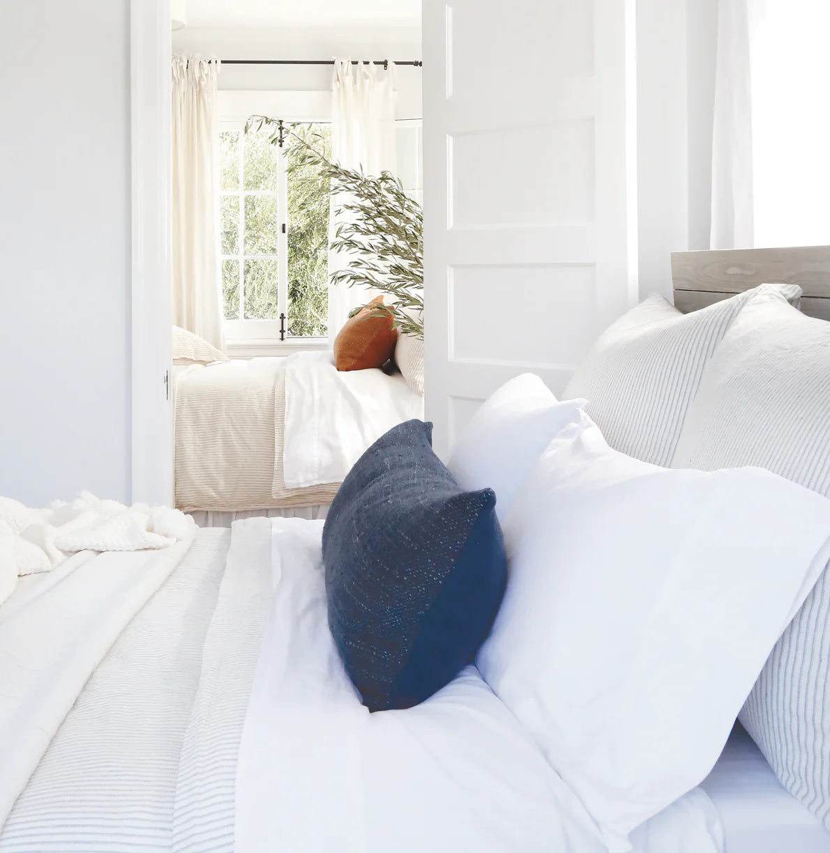 Bright, airy bedroom with white bedding and a navy blue, handwoven POM POM Hendrick Body Pillow in the foreground. Through an open door, another bed with white covers, a rust pillow, and a leafy plant by the window is visible.