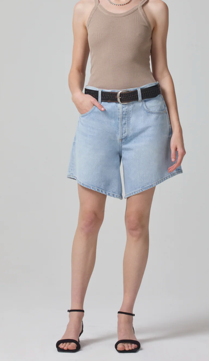 A person stands against a plain background, wearing a beige ribbed tank top, the Gaucho Skort in Dallas by Citizens of Humanity/AGOLDE made from high-rise light blue non-stretch organic cotton denim, a black belt, and black strappy sandals. Their hands are in their pockets as they face the camera.