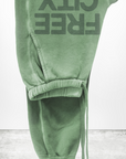 A pair of green FREECITY LARGE SUNFADES pocket sweatpant with the words "Free City (sparrow, LLC)" written in large block letters hang upside down against a white background. The lightweight French terry fabric appears soft and comfortable, featuring a drawstring waistband and elastic cuffs.