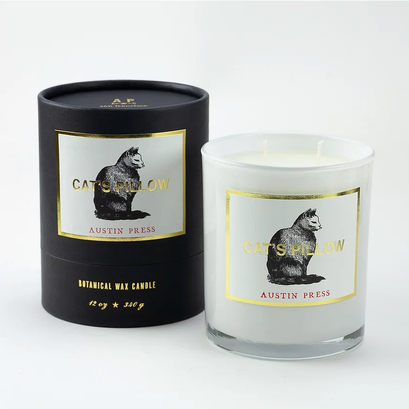 A Cat's Pillow Candle by Austin Press, labeled "Cat's Pillow," with a printed image of a cat on the label. The candle is beside its black packaging box, reminiscent of a cozy Scottsdale bungalow.