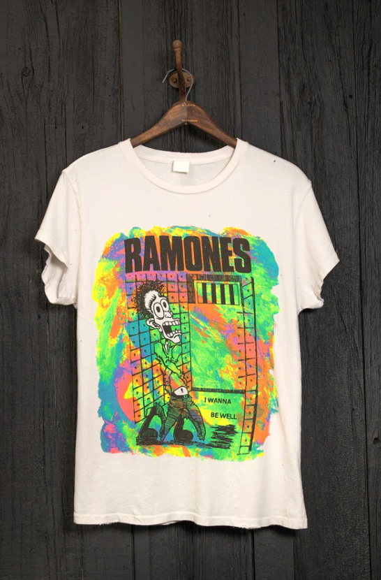 A white T-shirt with a colorful graphic of a skeleton and a rainbow-hued background. The text "RAMONES" is printed at the top in bold black letters, and below the graphic, it reads "I WANNA BE WELL." The T-shirt hangs on a wooden hanger against a dark wooden wall, evoking the laid-back vibe of Scottsdale bungalows. This is the RAMONES ESCAPE FROM NEW YORK VINTAGE WHITE by Made Worn.