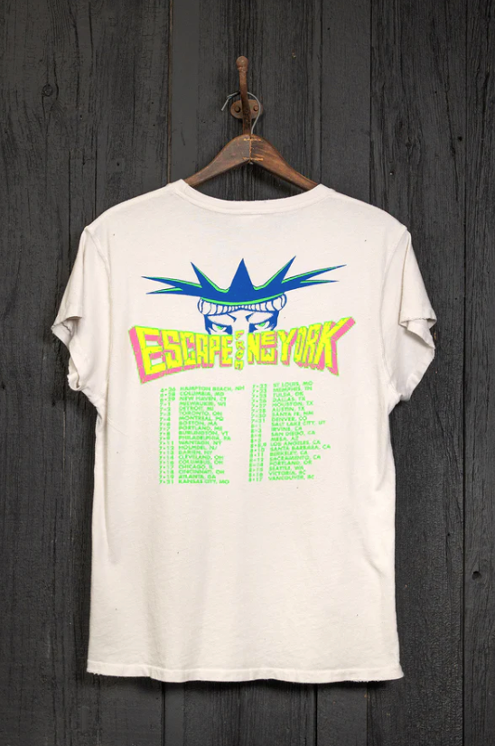 A RAMONES ESCAPE FROM NEW YORK VINTAGE WHITE hangs on a wooden hanger from a hook on a dark wooden wall. The back of the shirt features a vibrant blue and yellow graphic of the Statue of Liberty's crown with the words "Escape from New York" in bold font, reminiscent of souvenirs from Scottsdale, Arizona bungalows, and a list of text below.