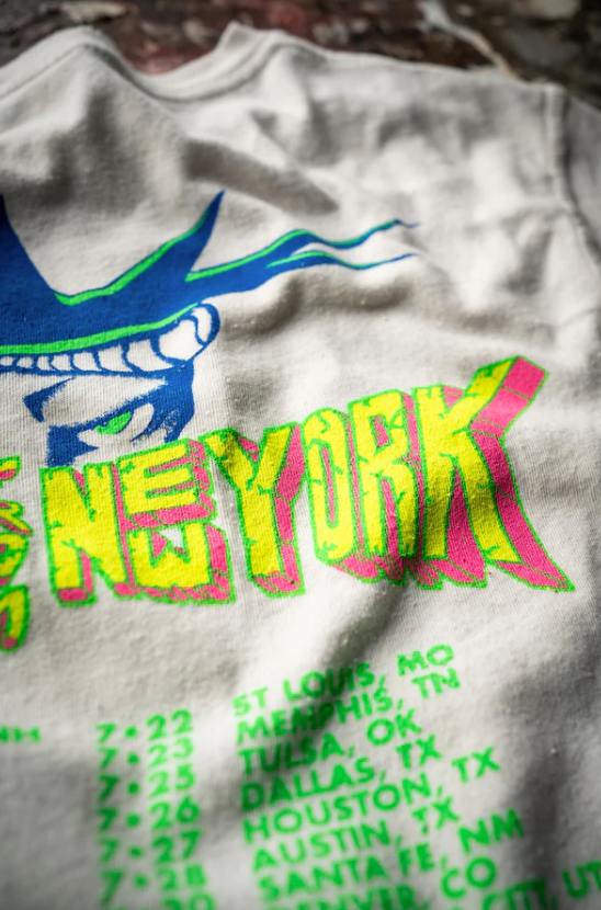 A close-up of a colorful, retro-style RAMONES ESCAPE FROM NEW YORK VINTAGE WHITE t-shirt by Made Worn featuring a graphic of the Statue of Liberty and the word "New York" in bold, neon letters. Below "New York," there is a list of cities and dates printed in green, including an iconic spot like Scottsdale, Arizona.
