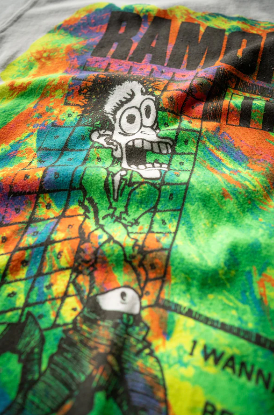 A colorful, tie-dye styled shirt featuring a cartoon character with a distressed expression. The text "RAMONES ESCAPE FROM NEW YORK VINTAGE WHITE" by Made Worn is partially visible at the top. Perfect for lounging in your Scottsdale, Arizona bungalow, the background includes vibrant hues of green, yellow, orange, and blue.