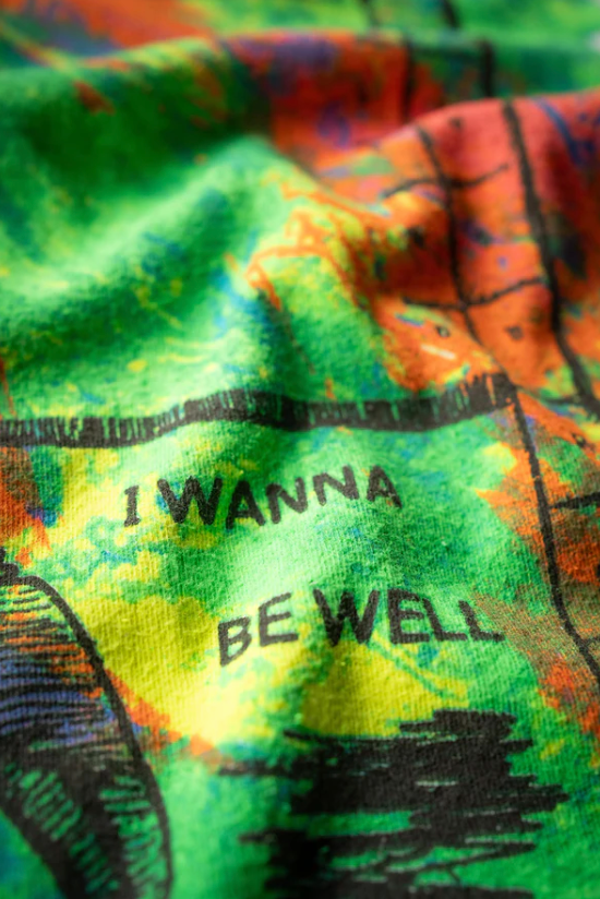 Close up photo of a colorful RAMONES ESCAPE FROM NEW YORK VINTAGE WHITE by Made Worn in a cozy Scottsdale, Arizona bungalow. The fabric has shades of green, yellow, red, blue, and orange, with the words "I WANNA BE WELL" printed in black on it.