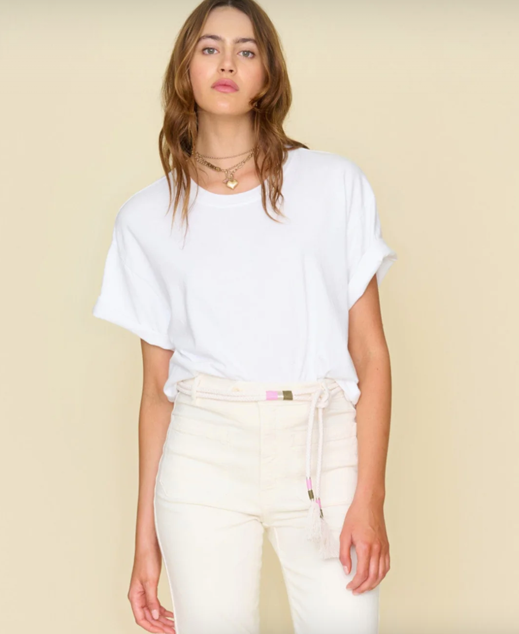 A person with long, wavy hair stands against a beige background reminiscent of a chic Scottsdale bungalow. They are wearing a loose-fitting White Palmer Tee by Xirena, tucked into high-waisted white pants. The outfit is accessorized with a belt and layered necklaces, exuding relaxed and confident vibes.