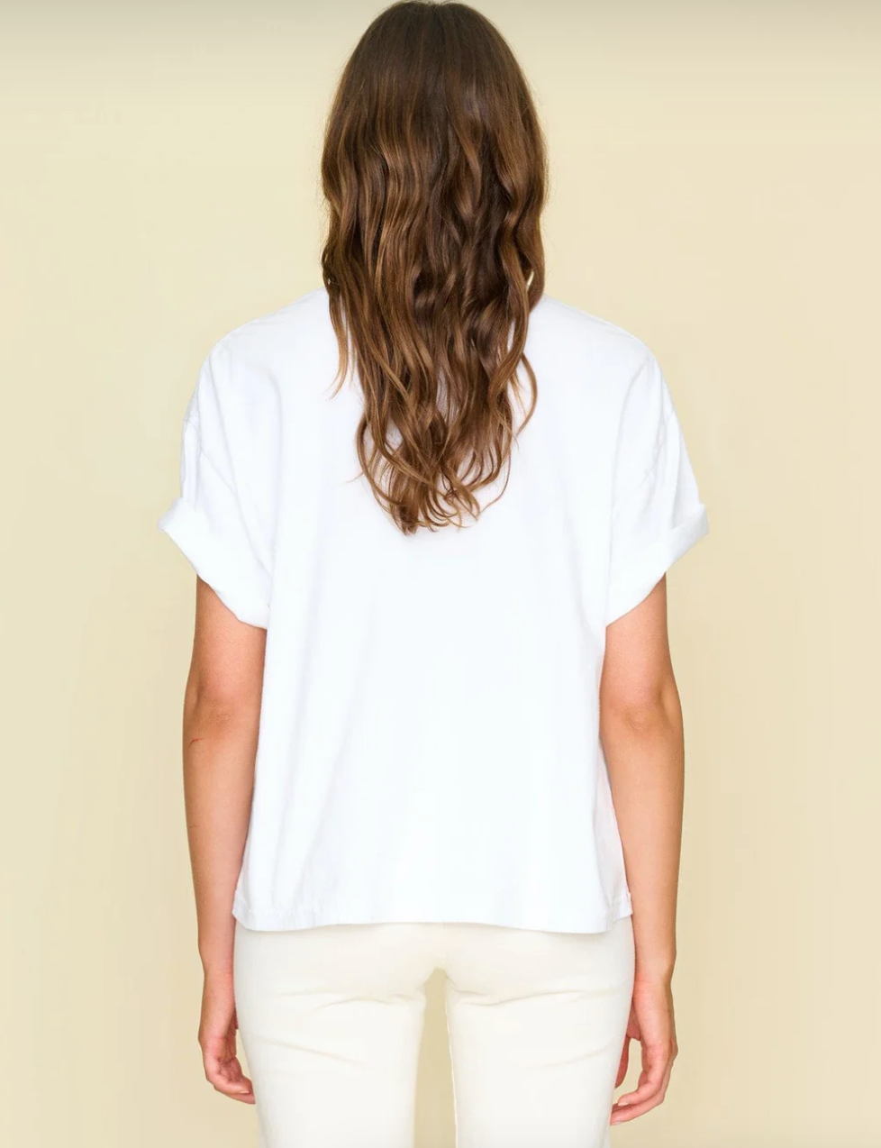 A person with long, wavy brown hair stands facing away from the camera. They are wearing a White Palmer Tee from Xirena and light-colored pants. The backdrop hints at minimalist elegance, reminiscent of a cozy bungalow in Scottsdale, Arizona, with its plain, light beige color.