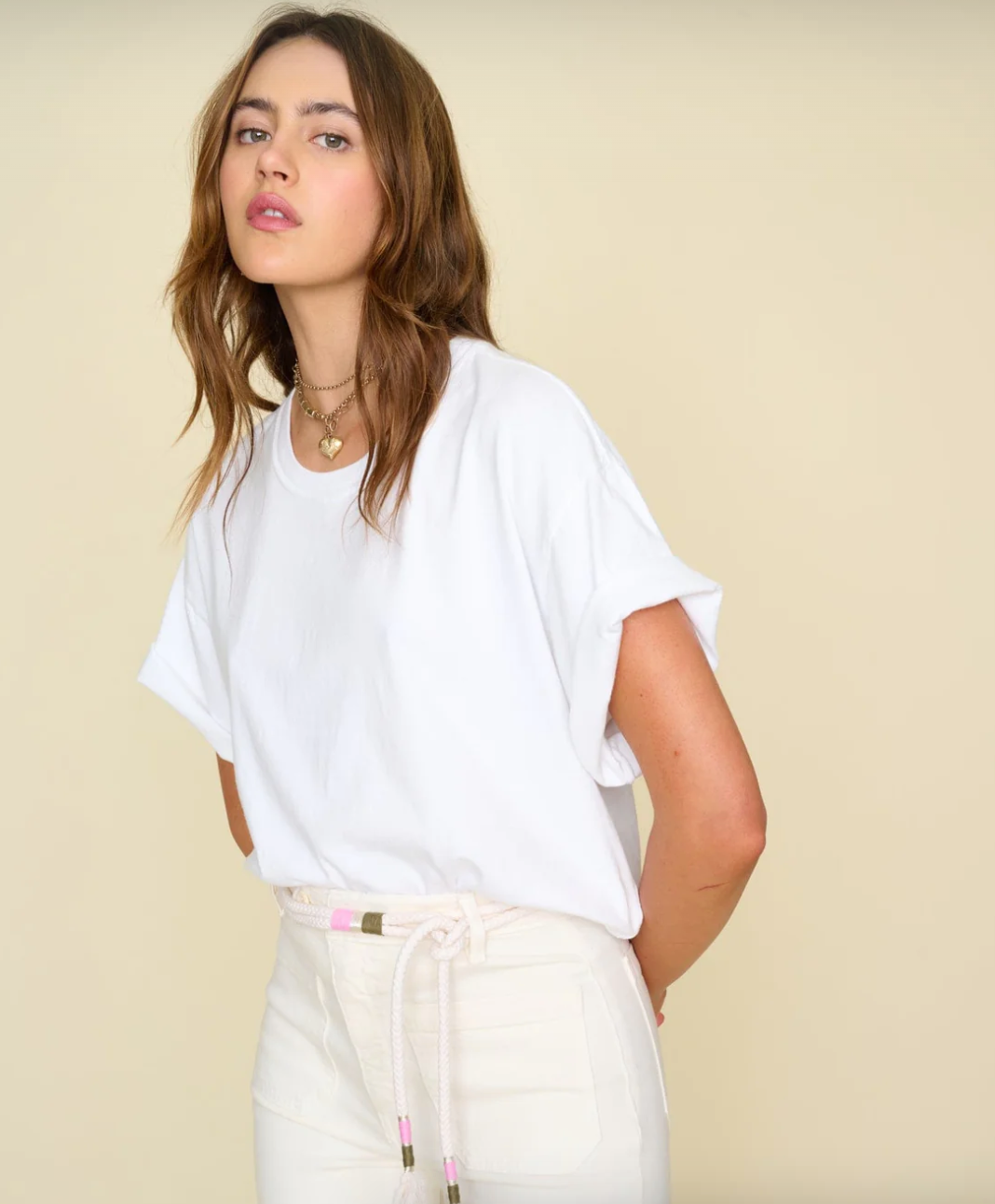 A person with long, wavy brown hair stands against a beige background, looking at the camera. They are wearing a Xirena White Palmer Tee and high-waisted white pants, with a decorative belt featuring beads and rope. With a relaxed, casual demeanor reminiscent of Scottsdale, Arizona bungalows, they exude effortless charm.