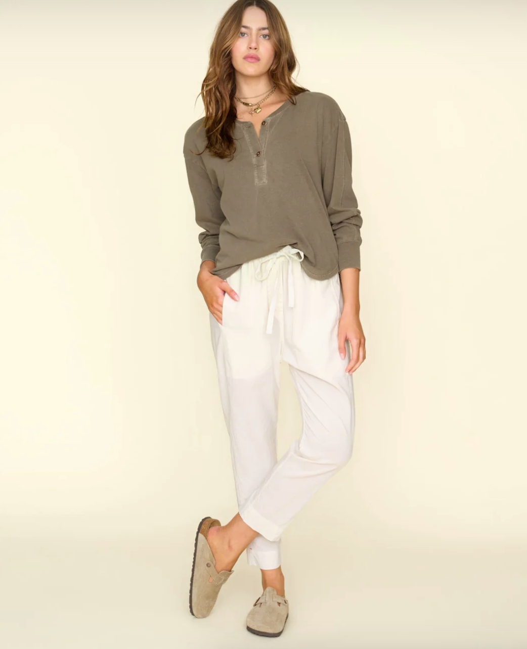 A person with long brown hair stands posing against a plain light background. They are wearing a sage green long-sleeved henley shirt, Xirena Sand Draper Pant, and beige slip-on shoes. With a relaxed posture and one hand in their pocket, they exude the effortless charm of a Scottsdale bungalow resident.