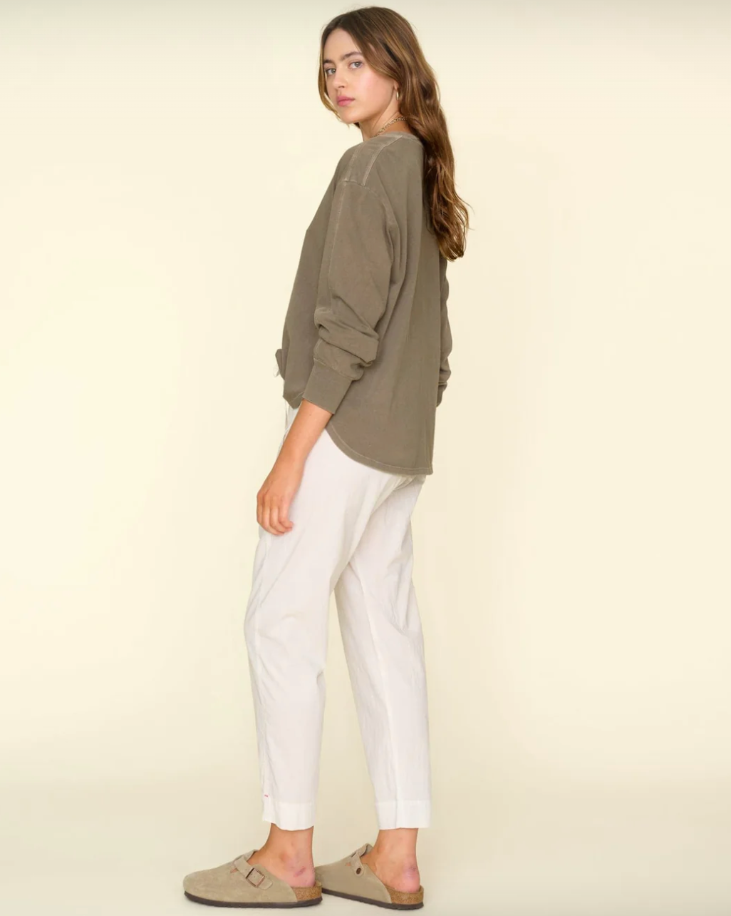 A person with long, wavy hair is standing sideways, facing left. They are wearing an oversized, olive-green sweater, Sand Draper Pant from Xirena, and beige slip-on sandals. The background is a solid light beige reminiscent of a cozy bungalow in Scottsdale Arizona. The person has a relaxed and casual demeanor.