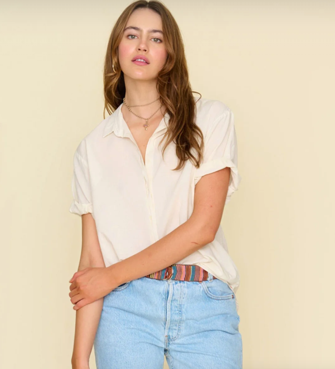A person with long brown hair stands in front of a beige background reminiscent of a Scottsdale, Arizona bungalow. They are wearing a light cream-colored Sand Channing Shirt by Xirena, slightly tucked into light blue jeans with a colorful striped belt peeking out. They have a calm expression and a relaxed posture.