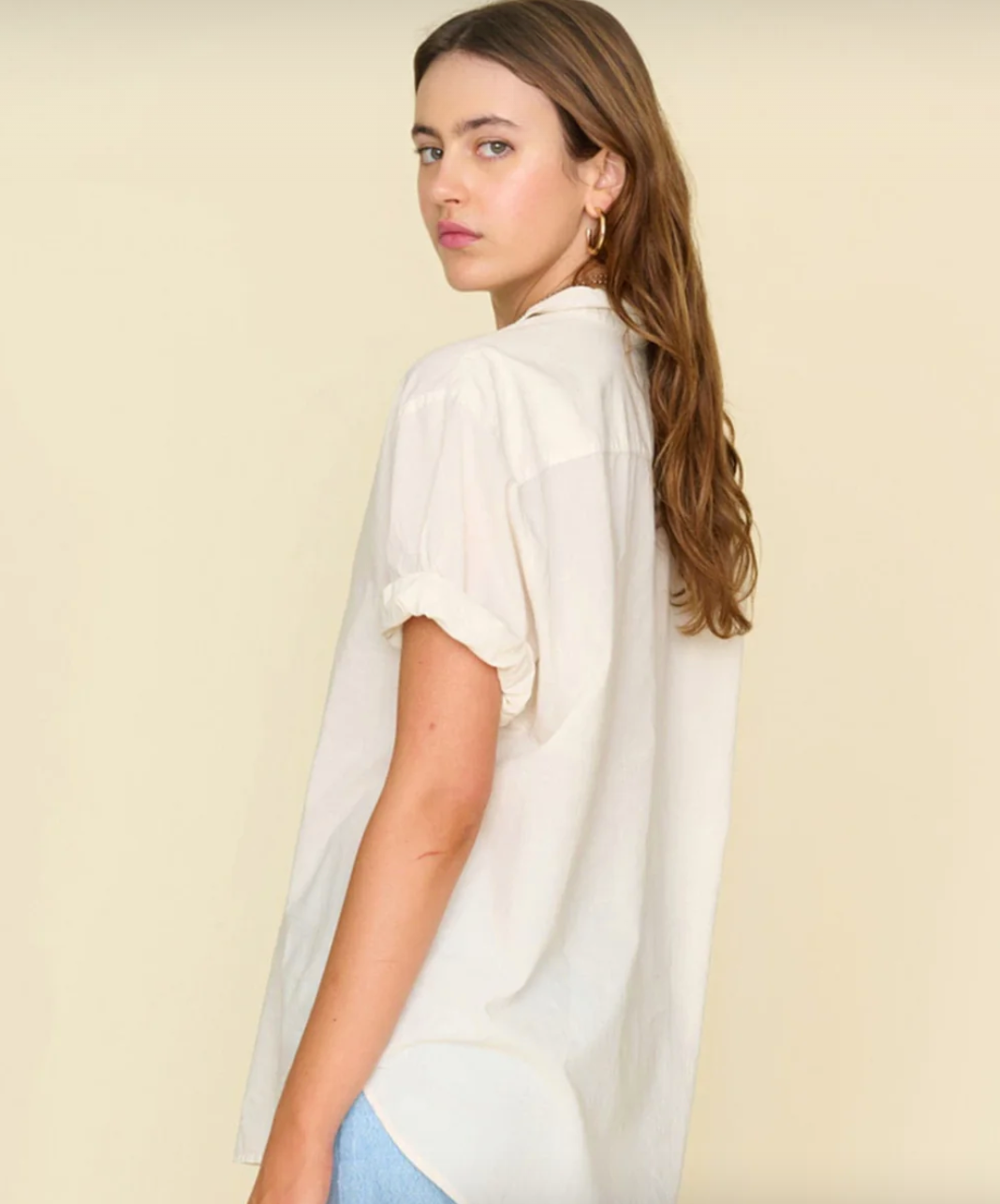 A person with long, brown hair is wearing a light-colored, short-sleeve Sand Channing Shirt by Xirena. They are standing against a beige background reminiscent of a Scottsdale Arizona bungalow, turned slightly to the side and looking over their shoulder at the camera with a neutral expression.