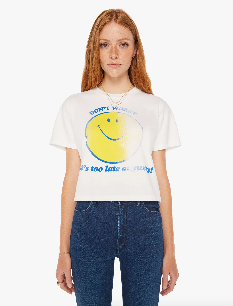 A woman with long red hair is wearing a white THE GRAB BAG CROP TEE DONT WORRY featuring a large yellow smiley face and the text "DON'T WORRY, It's too late anyway!" She is also wearing blue jeans. The background, reminiscent of a bungalow in Scottsdale, Arizona, is plain white.