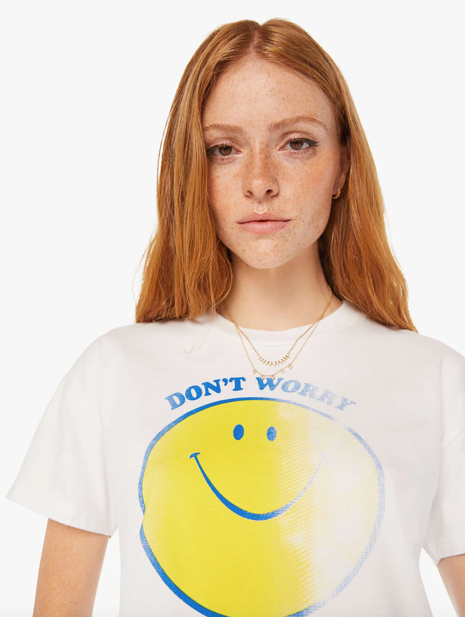 A person with long red hair and freckles wears a white t-shirt featuring a large yellow smiley face and the words "DON'T WORRY" in blue letters. They have a neutral expression and wear a layered gold necklace. This snapshot, reminiscent of relaxed Scottsdale Arizona vibes, has a plain white background, showcasing THE GRAB BAG CROP TEE DONT WORRY.