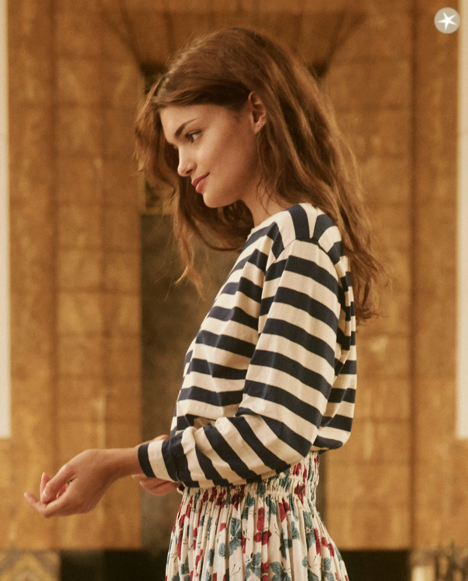 A young woman with long brown hair stands sideways, gazing downward with a serene expression. She wears The Great Inc.'s THE CAMPUS CREW NAVY AND CREAM SCHOLAR STRIPE and a colorful patterned skirt. The backdrop evokes the warm brown tones of a Scottsdale, Arizona bungalow, showcasing intricate architectural details.