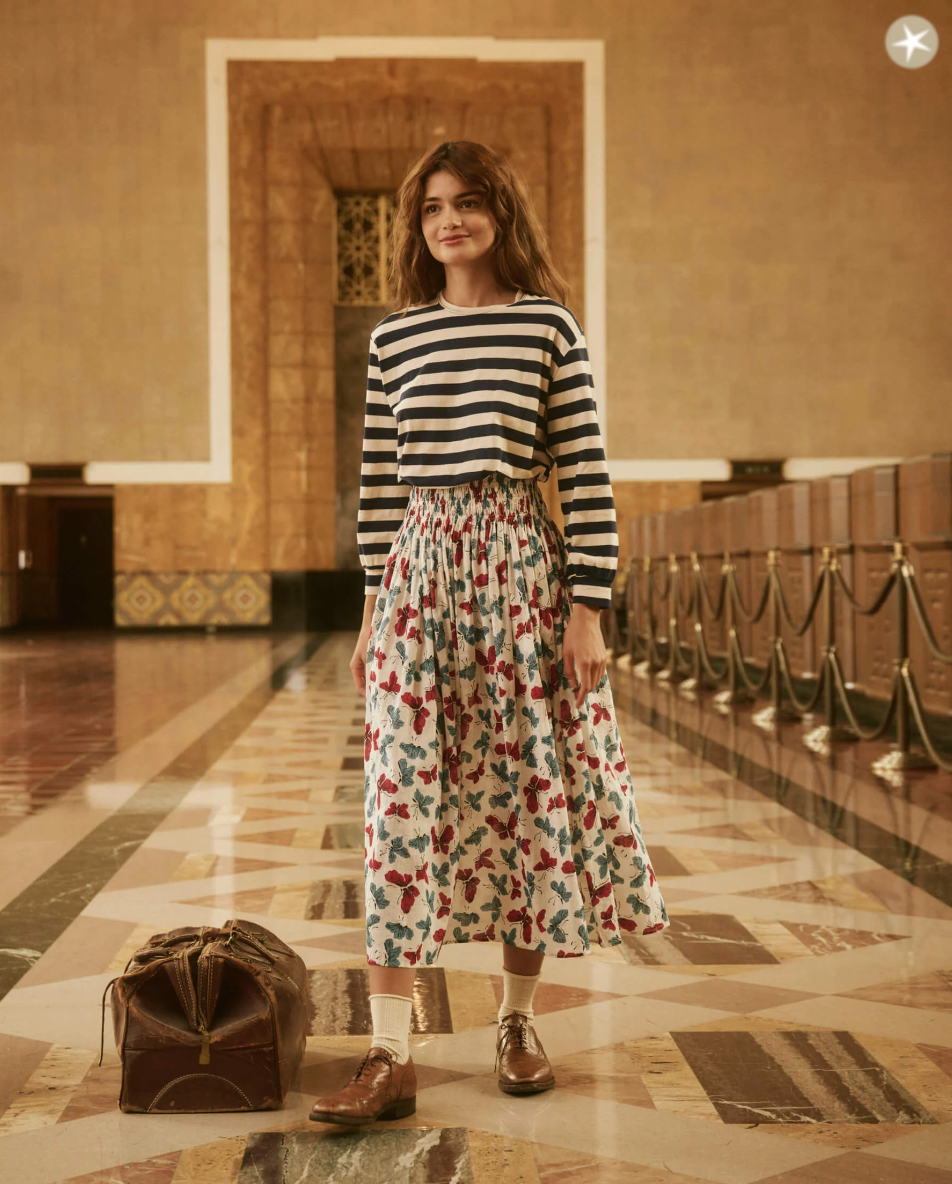 A woman stands in a spacious, grand building hall with high ceilings and patterned floors. She wears THE CAMPUS CREW NAVY AND CREAM SCHOLAR STRIPE by The Great Inc. and a floral long skirt paired with brown shoes. A vintage suitcase sits on the floor beside her, evoking the warm and classic ambiance reminiscent of a stylish bungalow in Scottsdale, Arizona.