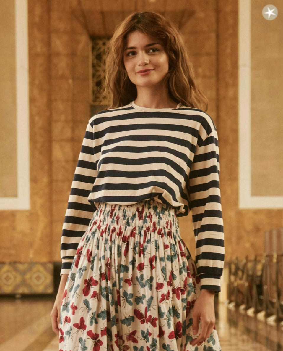 A woman with wavy brown hair stands indoors wearing a long-sleeved THE CAMPUS CREW NAVY AND CREAM SCHOLAR STRIPE by The Great Inc. paired with a high-waisted, floral, pleated skirt. The golden-hued interior features intricate architectural details reminiscent of a luxurious bungalow in Scottsdale, Arizona.
