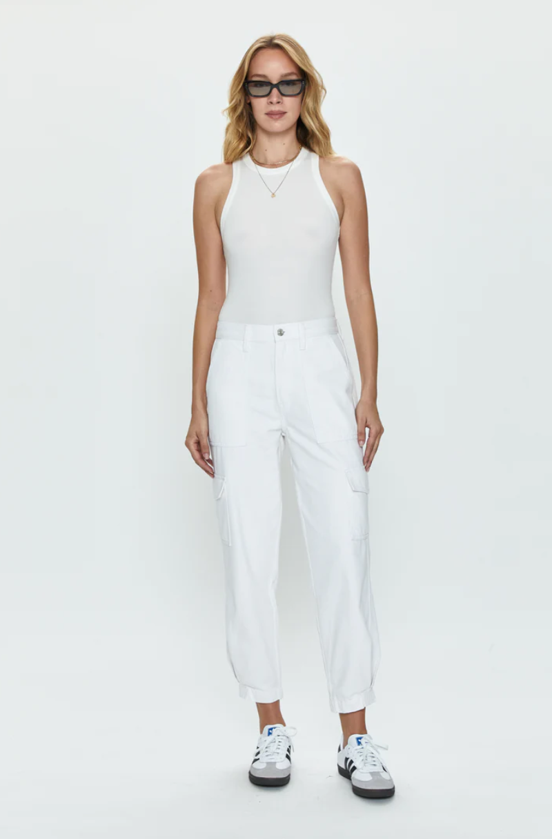 A woman stands confidently against a white background, wearing Pistola's Josephine High Rise Tapered Cargo in white, a Los Angeles-based white sleeveless top, white cargo pants, and white sneakers, accessorized with round sunglasses.