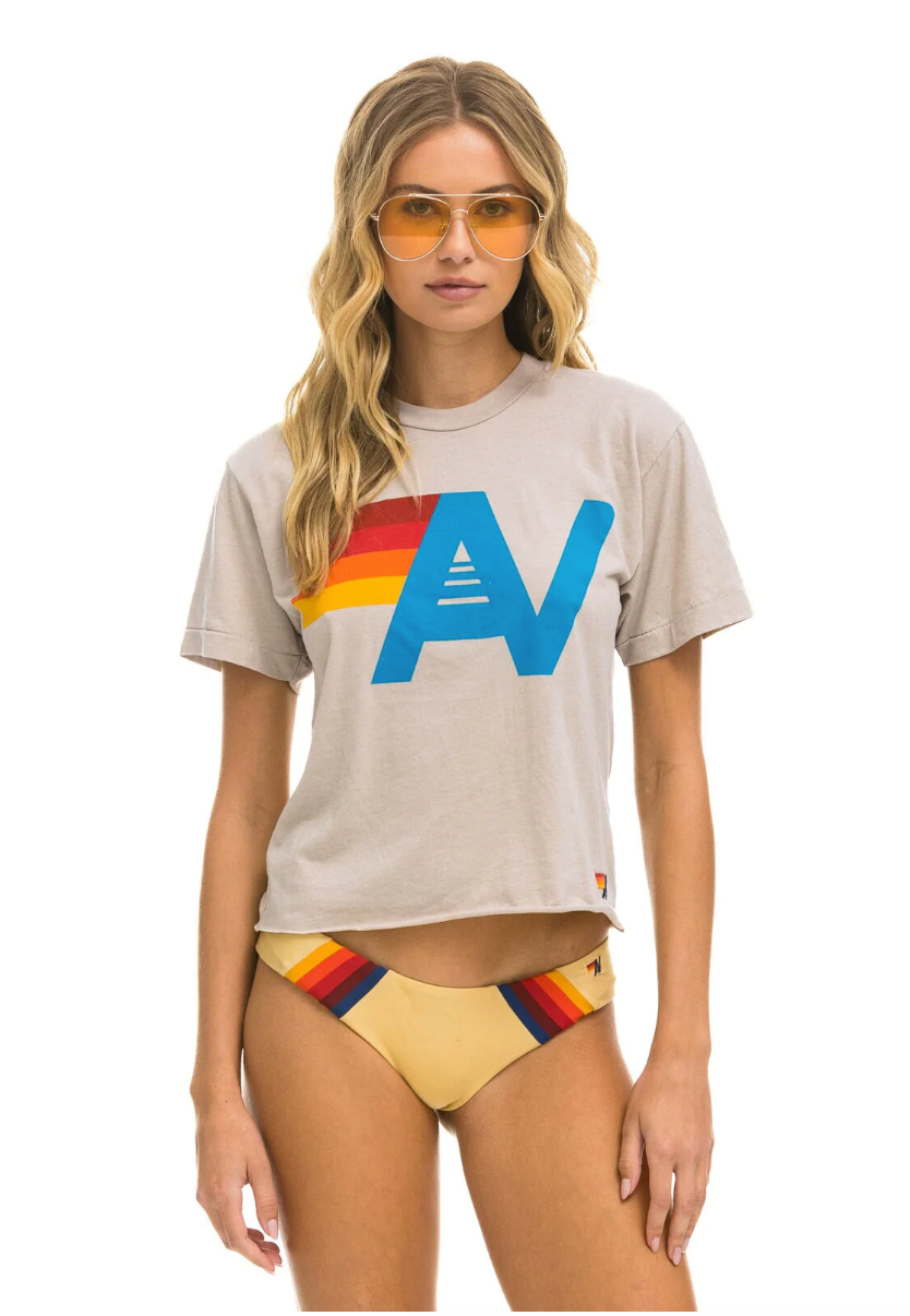 A woman with wavy blonde hair, wearing yellow-tinted sunglasses, a LOGO BOYFRIEND TEE - SAND by Aviator Nation  featuring a colorful "A" and stripe design, and beige bikini bottoms with matching red, yellow, and orange stripes stands against a white backdrop. Her look perfectly captures the laid-back vibe of Scottsdale, Arizona.