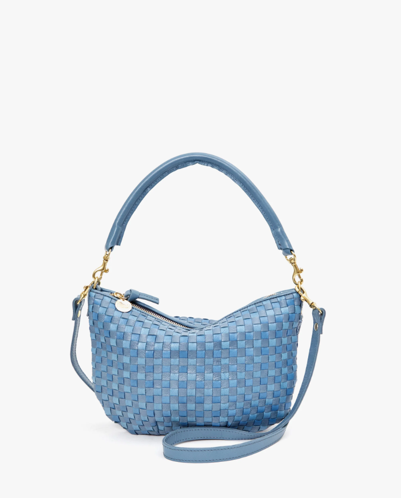 A small, blue handwoven leather handbag with a top handle and a detachable crossbody strap. This Petit Moyen Woven Checker mini messenger bag by Clare Vivier features a gold zip-top closure and gold hardware where the strap connects to the bag. The background is plain white.