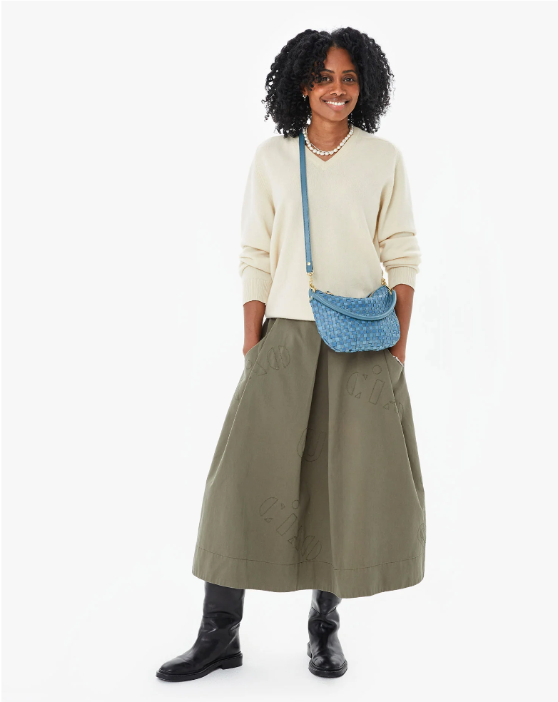 Person with curly hair, smiling and wearing a cream sweater, olive green maxi skirt, and black boots. They have a Clare Vivier Petit Moyen Woven Checker with a zip-top and a necklace. Background is plain white.