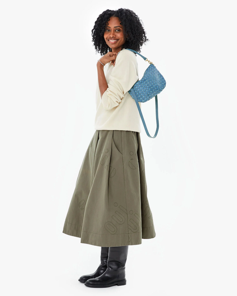 A woman with curly black hair stands smiling, wearing a cream sweater, olive green embroidered midi skirt, and black boots. She has a blue Petit Moyen Woven Checker mini messenger bag by Clare Vivier resting on her shoulder. The background is plain white.