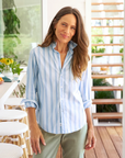 A woman with long brown hair smiles softly, wearing a Frank & Eileen BARRY Tailored Button-Up Shirt SUPERLUXE Slate Blue Stripe and green pants, standing in a brightly lit bungalow with plants in the background.