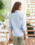 A woman stands facing away from the camera, wearing a Frank & Eileen BARRY Tailored Button-Up Shirt SUPERLUXE Slate Blue Stripe and green pants in a bright room with plants and a wooden deck visible through a large window in an Arizona-style bungalow.