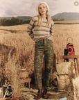A woman in The Shrunken Pullover. WATERFRONT STRIPE by The Great Inc. and camouflage pants stands in a golden wheat field with camping gear, a lantern, and a dog beside her, showcasing an Arizona style.
