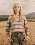 A woman in a striped sweater and camo pants stands in a wheat field, wearing a white beret styled as a nod to Arizona fashion, with distant mountains under a cloudy sky, The Shrunken Pullover from The Great Inc.