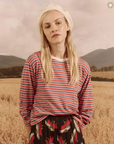 A woman wearing The Campus Crew CAMPERVAN STRIPE shirt and a floral skirt stands in a wheat field, with mountains in the background and a clear Arizona sky. She has a thoughtful expression and wears a white beret.