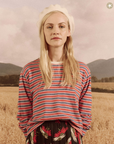 A woman with blond hair sporting a white beret and a striped long-sleeve shirt stands in front of a pastoral field with distant mountains under a subdued Arizona sky wearing The Great Inc.'s The Campus Crew CAMPERVAN STRIPE.
