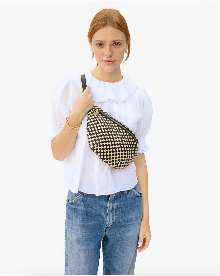 A woman with red hair wearing a white blouse and Arizona-style blue jeans holds a Clare Vivier Grande Fanny Woven Checker shoulder bag. She stands against a plain background.